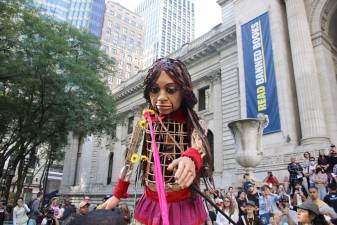 Little Amal in front of the New York Public Library, reaching towards flowers presented to her. Photo: Gaby Messino