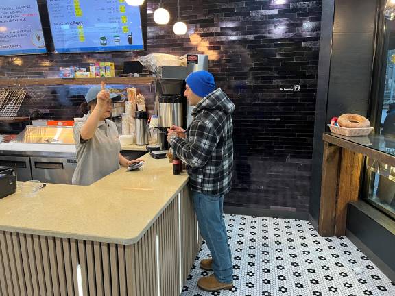 Manager Amy (behind counter) greets the first customer to New Absolute Bagels at the pickup area; as he waited for his bagel with topping. The ,man made history at 8:15 a.m. as the first customer at the just-opened store. It didn’t take long for this space to fill up thereafter.