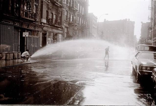 On East Fourth Street, one man tries to block an exploding fire hydrant while another runs under the arc of water (1970). Photo courtesy of Alex Harsley